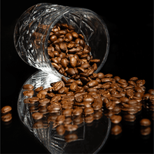 Glass with spilled Bourbon Barrel Aged Coffee beans on a reflective surface, showcasing their rich color and texture.