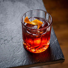 A crystal bourbon glass filled with a vibrant cocktail, garnished with an orange twist, resting on a dark wood table.